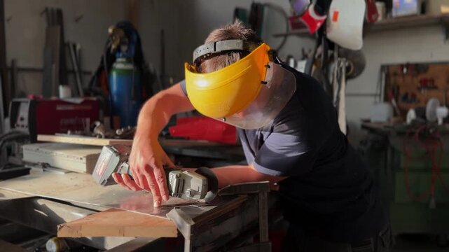 Focused artisan wearing a protective face shield meticulously cutting a metal sheet with a powerful angle grinder in a busy workshop