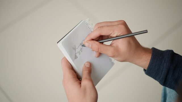 Close up of hands drawing sketch in notebook with pencil capturing detailed line art progress on white paper against plain background. Hands hold spiral bound notepad steady while right hand