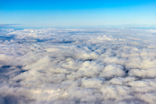 An aerial view of a thick layer of fluffy white clouds under a clear blue sky,  captured from an airplane window at high. A dense, rolling blanket of  clouds filling the lower portion of the frame.