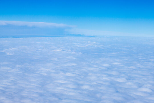 An aerial view from an airplane window looking out over a thick, rolling layer of low-level clouds beneath a clear blue sky. A vast sheet of white, textured clouds, typical of a low cloud layer. 