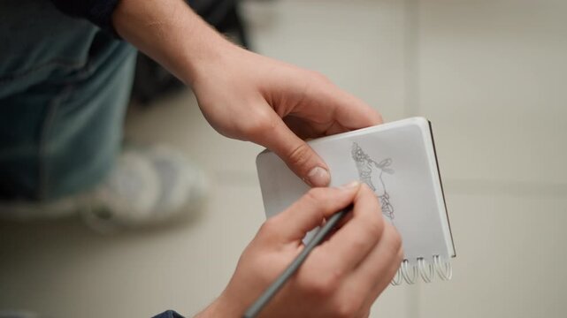 Close up of hands drawing sketch in spiral bound notebook capturing detailed pencil work on paper. Hands hold book steady while adding fine lines to existing illustration showing focused