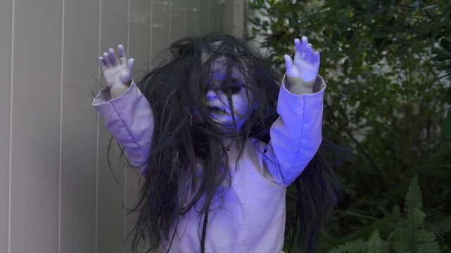 Spooky doll prop with a pale face, messy black hair, and raised hands in a yard. Halloween outdoor decoration in a garden with green plants creating a horror mood.