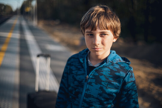 A young boy in a blue hoodie standing with a suitcase on an outdoor train station platform, waiting for a train.