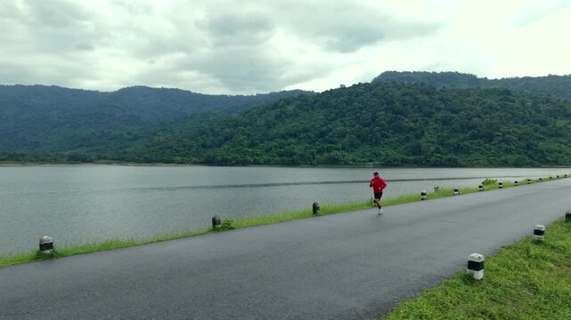 Drone view of a man running along a dam ridge during an outdoor workout. The scene represents fitness goals, personal challenge, determination, achievement and an active healthy lifestyle.
