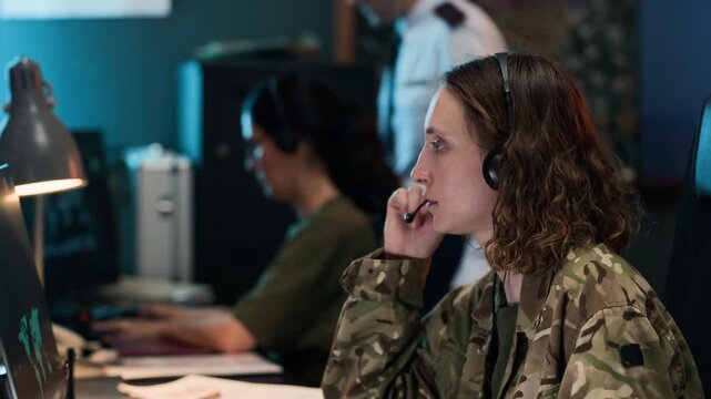 Young adult Caucasian female soldier wearing headset typing on computer in military command center. Clip is suitable for defense technology, cybersecurity, and national security marketing