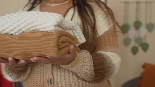 Woman holds folded knit sweaters and places them on a white closet shelf with visible hand in a small dressing area building; tidying organization calm.