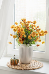 Naklejka premium Orange chrysanthemum bouquet in white vase with small pumpkin on bright windowsill, seasonal autumn home decor with natural daylight. Vertical format. Cozy home interior