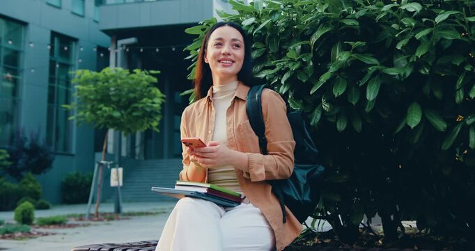 Smiling professional businesswoman typing on a smartphone outdoors near a modern office building. Remote work concept, digital communication, corporate lifestyle, urban business environment.