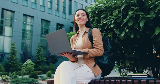Concentrated young woman freelancer sitting on bench typing on laptop of background modern building. Serious pensive girl doing remote job. Outdoors