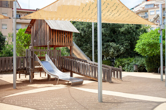 Wooden playground structure with metal slide under large shade sails in a public urban park. Outdoor recreational area for children with safe play equipment, natural materials and sunny day atmosphere