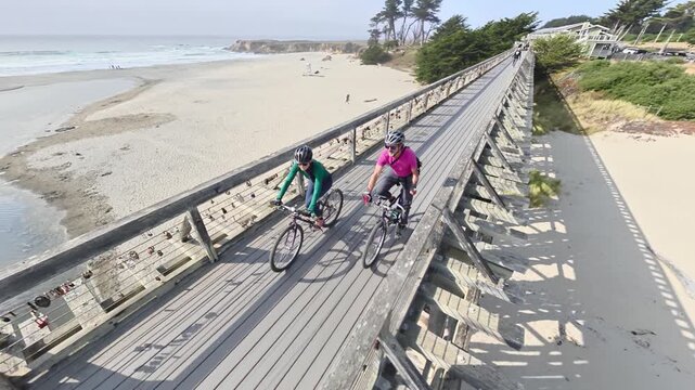 Senior man and woman ride bikes together on a bridge next to the ocean in Fort Bragg, California enjoying the sunny weather.