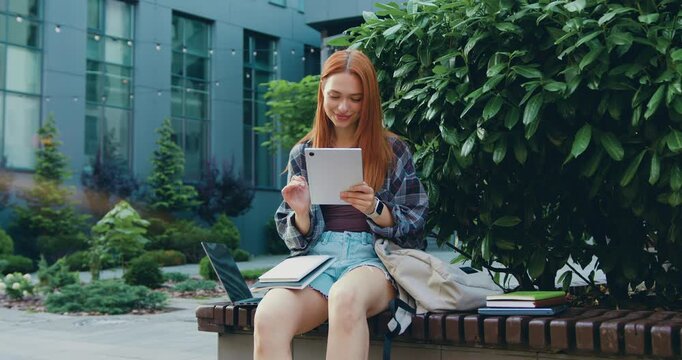 Close-up of beautiful girl smiling while using tablet in urban park during sunny day surrounded by commercial glass buildings in background