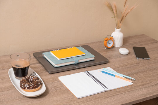 Desk setup with coffee, donut, notebooks, and clock during midday