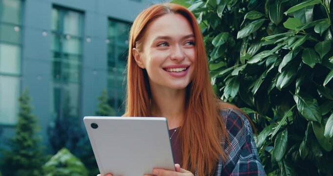 Joyful student woman on the street while holding a digital pad in front of her, engaged in an online studying. City buildings visible in the background.