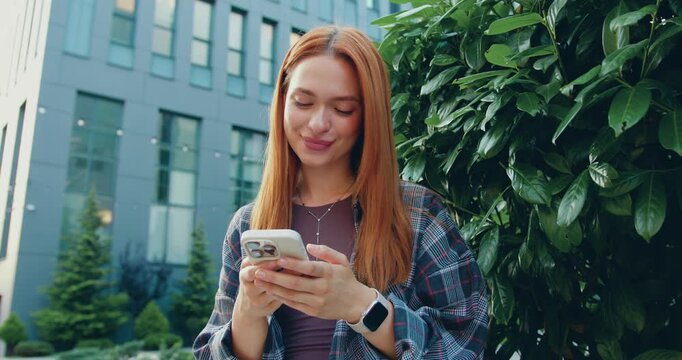 Face portrait of brunette woman holding smartphone, scrolling social media texting browsing online while wind blowing at her hair against the background of modern building