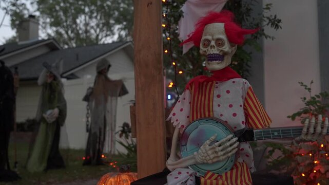Close-up of a skeleton clown with glowing red eyes playing a banjo at dusk. Orange lights, a pumpkin, and witches in the background create a spooky Halloween yard decoration.