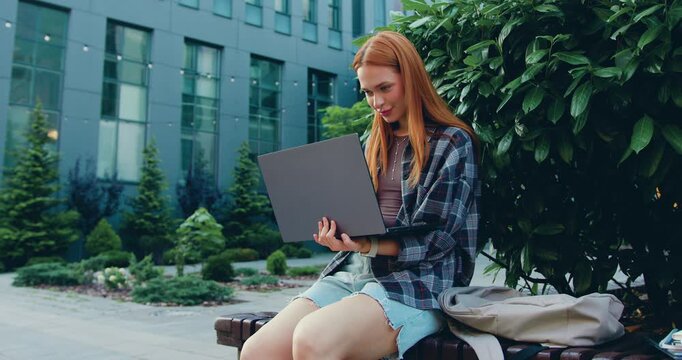 Smiling young woman typing laptop sitting urban bench near modern building. Focused female freelance working remotely on street in modern park