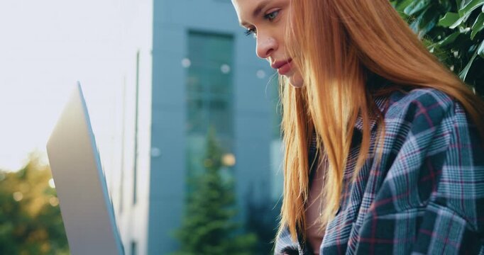 Close up. Portrait of a young woman working alone on a laptop outside, looking at the screen with focus and friendliness while performing an online remote job, with a glass office building in the