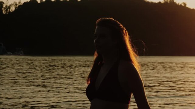 Woman walks along the beach at dusk her silhouette framed by fading sunlight as the ocean stretches out to horizon. In graceful slow motion she pauses for portrait looking towards camera with smile