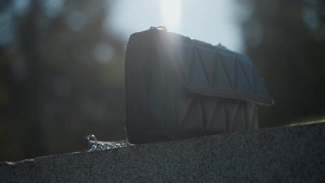 Close up of black handbag resting on stone with sunlight backlighting. bag remains stationary throughout showcasing its texture and geometric design against outdoor background with soft