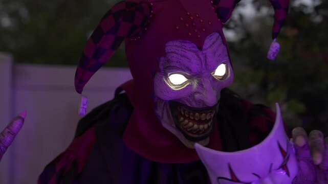Close-up of a scary Halloween jester clown with glowing eyes at night. The figure holds a white mask, showing a creepy evil smile as an outdoor yard decoration.