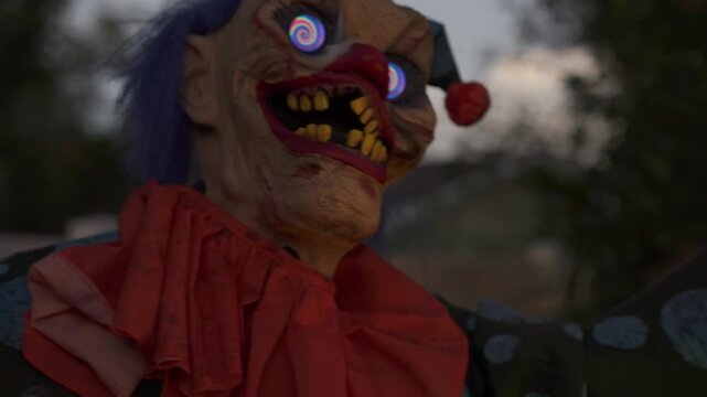 Close-up of a scary Halloween clown with spiral glowing eyes, green face, rotten yellow teeth, blue hair, and a red nose at night. Outdoor yard decoration with an orange collar creates a spooky horror