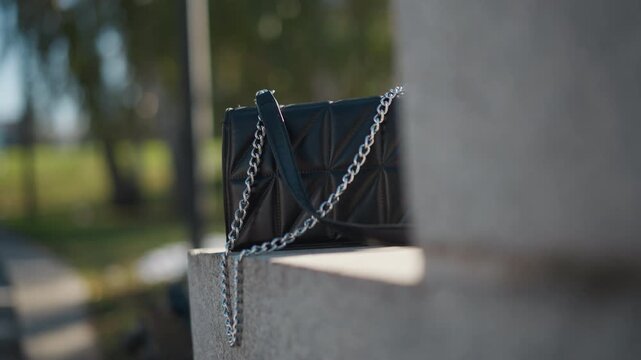 Close up of black leather handbag with silver chain strap resting on concrete in outdoor park setting. Bag remains stationary throughout showcasing quilted texture and sleek design