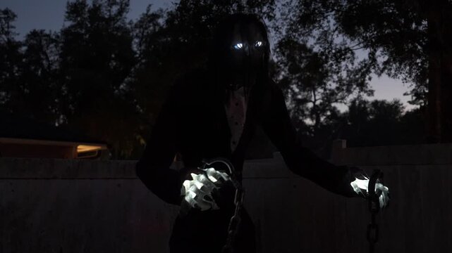 Dark Halloween figure with glowing eyes and bright hands holding metal shackles by a fence at dusk. Spooky outdoor yard decoration creates a scary night horror scene.