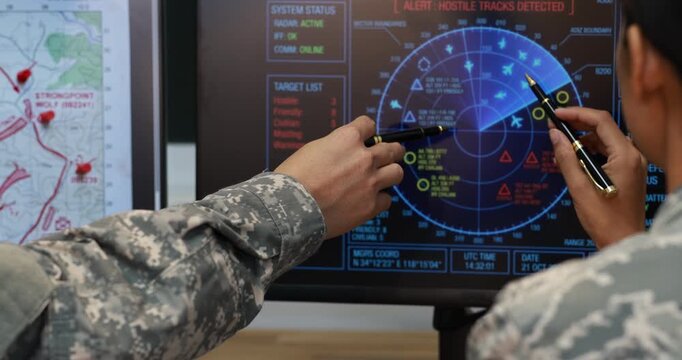 Military personnel point at a digital radar screen an alert and map data during a command center briefing.