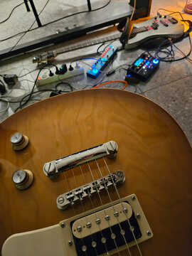 Guitar with vintage finish and chrome hardware. Strings rest taut over bridge and pickups. Pedals and cables lie scattered nearby. Warm wood grain glows under soft lighting