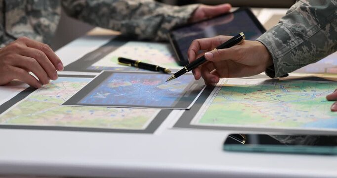 Military personnel in camouflage uniforms review printed maps and point with pens during a planning meeting at a table.