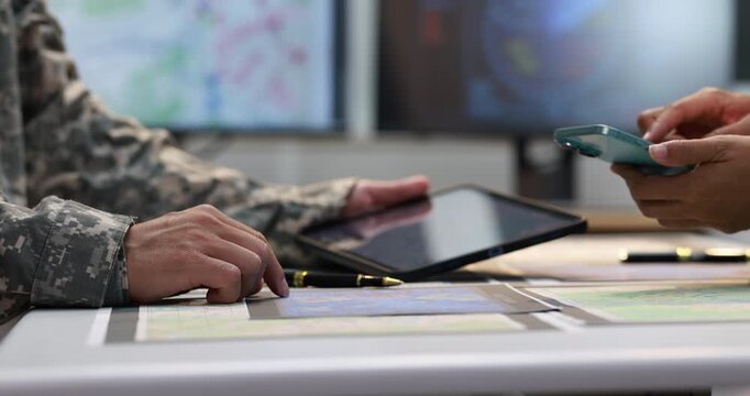 Close-up of a military planning meeting with a camouflage-clad person pointing at a map while another uses a smartphone beside a tablet and documents.