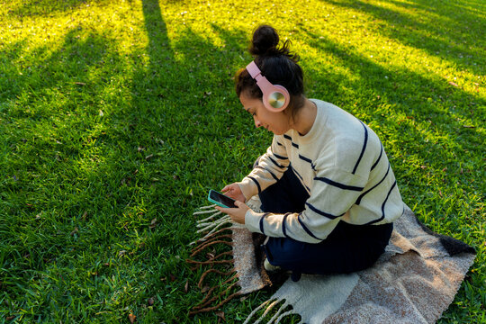 Woman enjoying music in park, using smartphone and wearing headphones, relaxing on grass outdoors