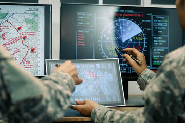 Military personnel review tactical maps and tracking screens in a command center, using digital displays and a tablet for mission planning and battlefield coordination. © Phushutter