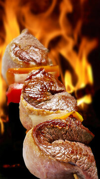 Close up of juicy Brazilian picanha beef skewers with peppers roasting over an open fire. Traditional barbecue steak sizzling against bright flames.