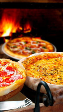 A close-up shot of two delicious freshly-made artisan pizzas (pepperoni and a cheese with herbs) with a roaring wood-fired oven glowing in the rustic background.