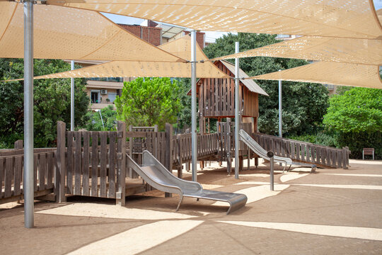 Wooden playground structure with metal slide under large shade sails in a public urban park in Barcelona, Spain. Outdoor recreational area for children with safe play equipment, natural materials 