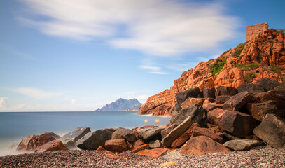 Genoese tower above Porto Beach on Corsica, with red granite cliffs, dark coastal boulders and silky long-exposure sea beneath drifting clouds. © Petr