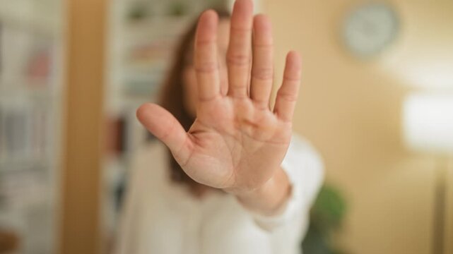 Woman holds palm forward in stop gesture toward camera in a home studio with bookshelves, clock and lamp visible; privacy defiance.