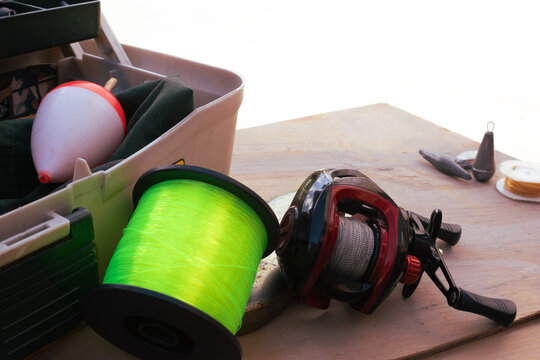 Fishing gear arranged on a wooden surface, featuring a baitcasting reel, bright green fishing line spool, tackle box, bobber, and lead weights, preparing for an outdoor recreational activity
