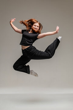 Teenage girl jumping in air with flying hair in black clothes. Young dancer performing dance move on gray studio background. Joy, energy and motion concept