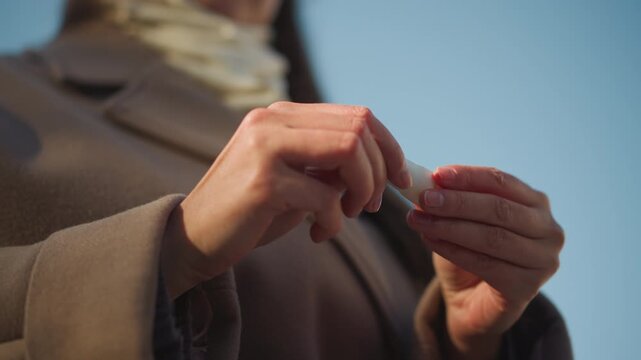 Close up of woman holding and opening small white bottle outdoors under clear blue sky. Hands with painted nails manipulate object emphasizing interaction between hands and product