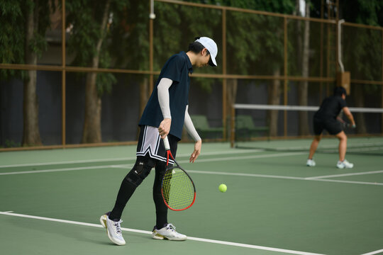 Asian Tennis Player Bouncing Ball on Outdoor Green Court Before Serve with Opponent in Background