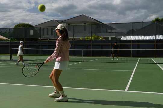 A young woman in pink jacket watches a tennis ball fly out of bounds during a doubles competition on a green outdoor court.