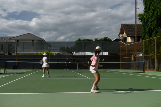 Two Women Playing Tennis Doubles Match on Outdoor Green Court with Cloudy Sky Background