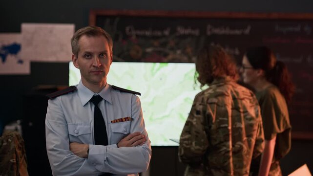 Portrait of middle-aged Caucasian male military officer standing with arms crossed in command center while colleagues analyze map, zooming in. Footage is ideal for defense industry marketing