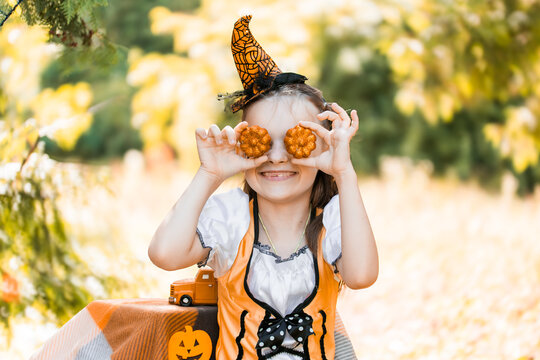Girl Dressed for Halloween Holds Pumpkin-Shaped Cookies in Front of Her Face in an Autumn Setting With Colorful Leaves in October