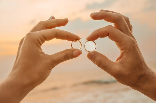 Couple holds wedding rings together during sunset by ocean. Two hands hold wedding rings while standing near ocean. sun sets in background,  warm glow. scene captures  moment of love commitment