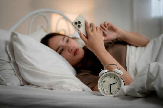Insomnia concept of a young woman using a smartphone while lying awake in bed at night beside an alarm clock, showing sleeplessness, insomnia, smartphone, bedtime, sleep, fatigue	