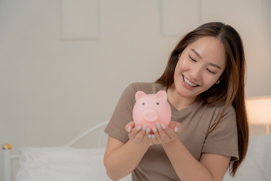 Saving money, financial planning and future goals concept. Happy young Asian woman holds a pink piggy bank at home. Savings, finance, investment, budget, future